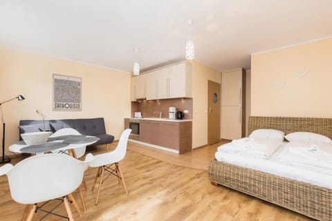 The room features a queen-size bed in an open-plan space. There’s a round dining table with two modern chairs, a compact, sleek design, and a TV mounted on the wall.