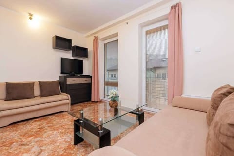  A cozy living room area with a beige couch, glass coffee table, and a large window that lets in natural light. Beige curtains and a wall-mounted TV are visible.