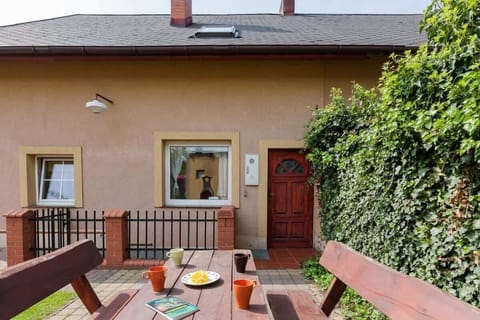View of the house from the front. A wooden table with benches is visible in the foreground and the entrance to the house can be seen behind them. 