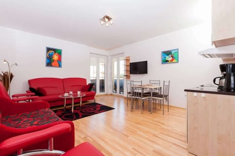 Spacious living room with wooden flooring. In the centre is a large red sofa and a glass coffee table. In the background you can see part of the kitchen with white cabinets and a dining table with several chairs.