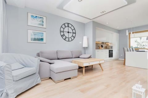 A bright living room featuring a cozy gray sofa, a modern coffee table, large windows with outdoor views, and stylish decor including a large wall clock.