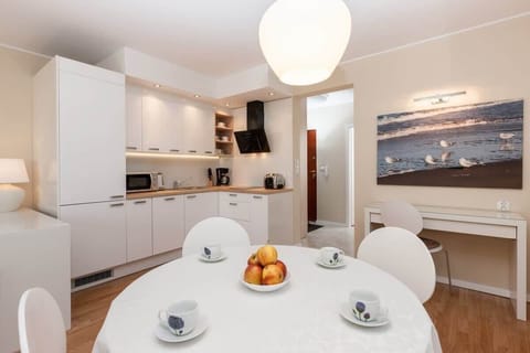 A sleek, modern kitchenette with white cabinets, integrated appliances, and a dining table in the center. The table is set with white chairs and is illuminated by a stylish hanging light.