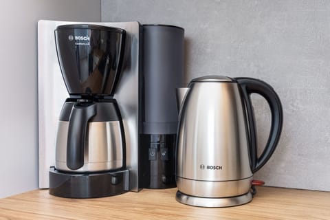 A close-up of a modern coffee station featuring a coffee machine, electric kettle, and cups, placed on a wooden counter for a homey touch.