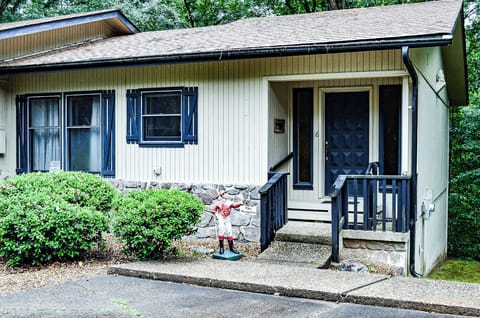This charming cottage exterior welcomes travelers with classic shuttered windows and a friendly entrance porch surrounded by lush greenery and mature trees, creating a peaceful woodland retreat setting.