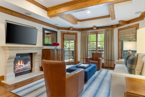 Living room with leather armchairs, blue tufted ottoman, stone fireplace, and wood-trimmed windows opening to a balcony.