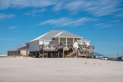 Beachfront vacation home elevated on stilts with wraparound decks, situated directly on pristine white sand under bright blue skies.