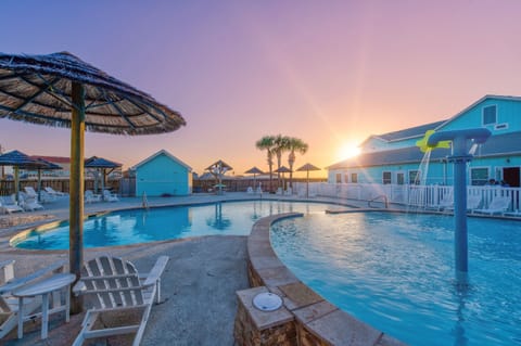Beachside retreat featuring colorful cottages, sparkling pool, and thatched umbrellas beneath stunning sunset skies.