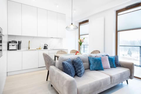 A spacious, modern living room featuring a large sofa adorned with blue cushions. Large windows bring in ample natural light.