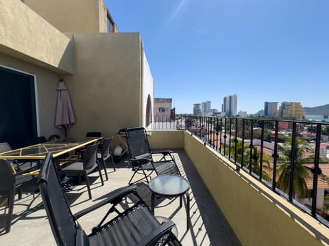 Spacious balcony with black metal railing, glass-top dining table, lounge chairs, and city skyline views.