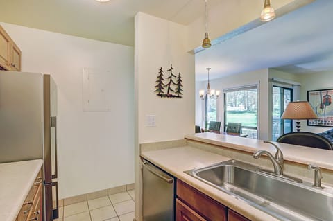 A kitchen area with a stainless steel sink, wooden cabinets, tiled floor, and minimalistic wall decorations, including tree-shaped art. The space opens to a dining area with a view of a grassy outdoor area.