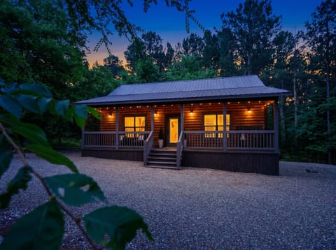 A charming log cabin nestled in the woods, glowing warmly under twilight skies with string lights and a welcoming porch. The property offers peaceful seclusion surrounded by nature.