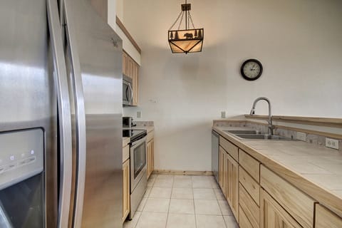 A modern kitchen with stainless steel refrigerator, oven, microwave, light-colored wooden cabinets, tiled countertops and floors, a clock on the wall, and a ceiling light featuring a bear design.