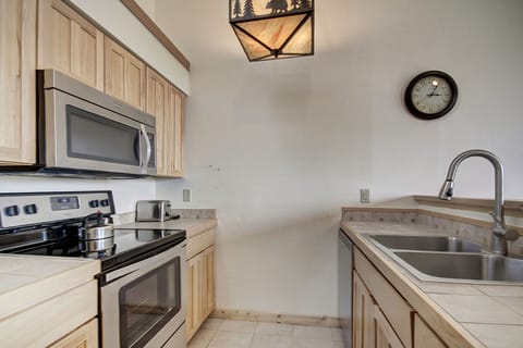 A modern kitchen featuring wooden cabinetry, a stainless steel stove with a pot on it, a microwave, a sink with a faucet, and a wall clock. The light fixture has a forest and wildlife design.