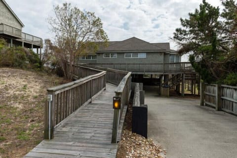 A raised beach home nestled among coastal dunes and pines, with elevated walkways leading to covered parking and the main entrance.