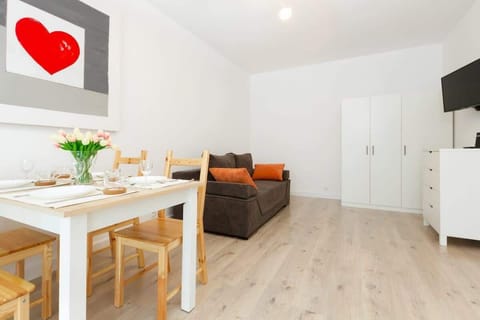 A bright, modern living room featuring a dining table with wooden chairs, and a comfortable sofa in the background. The decor is minimalistic with light wooden floors and white walls.