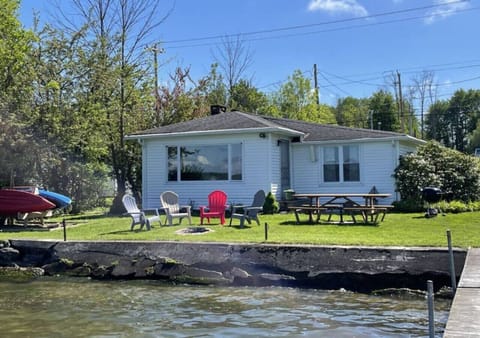 View of lawn ares in front of white cottage with kayaks, fire-pit, lawn chairs BBQ and picnic table.