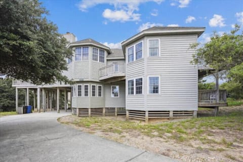 A spacious two-story coastal vacation home featuring classic beachside architecture with multiple balconies and elevated design, surrounded by mature coastal trees and offering covered parking beneath.
