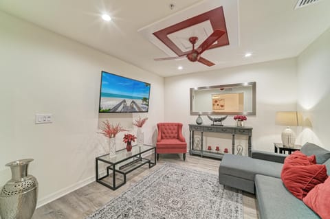 Living area with gray sectional sofa, red accent pillows, tufted armchair, glass coffee table, and wall-mounted TV.