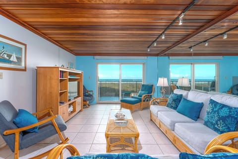 This incredible living room, adorned with mirrored walls reflecting the stunning beachfront view, features wicker furniture for 8 people, a flat-screen TV, and an inviting, sunny ambiance.