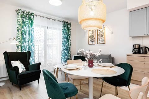 A cozy living room featuring a round dining table, green velvet chairs, and a large window with patterned green curtains. The space is bright and stylish.