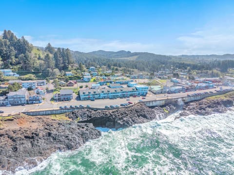 Aerial view of coastal town nestled between forested hills and dramatic rocky shoreline with crashing waves.