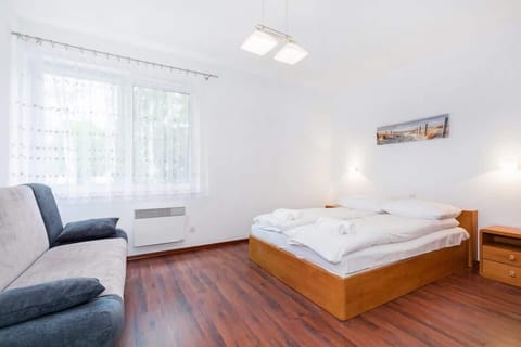 A bright bedroom with a wooden double bed, bedside tables, and a large window allowing natural light. The bed has white bedding and a simple artwork above it.