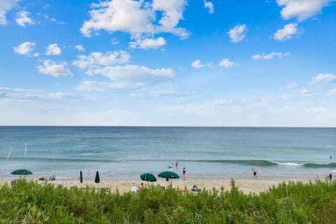 A pristine beach scene with gentle waves rolling ashore, umbrellas dotting the sand, and natural dune grasses swaying in the coastal breeze under a beautiful blue sky.