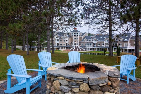 Outdoor fire pit with Adirondack chairs and greenery.
