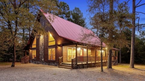 A welcoming log cabin with a distinctive red metal roof nestled among tall pines, offering a cozy woodland retreat with warm glowing windows and a spacious front porch.