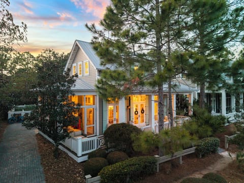 Charming coastal cottage with warm lighting glowing through French doors at dusk, surrounded by mature trees and manicured gardens.