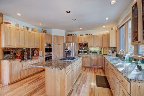 Spacious kitchen with wooden cabinets, granite countertops, stainless steel appliances, and hardwood flooring. Central island with cooktop and overhead lighting. Large window providing natural light.