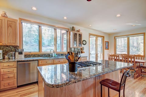 A spacious kitchen with wooden cabinets, granite countertops, a central island with a stove, and large windows providing natural light. A dining area with wooden furniture is visible in the background.