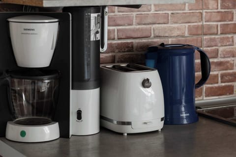  A coffee station with a coffee maker, a kettle, and cups arranged on a countertop against a brick wall.