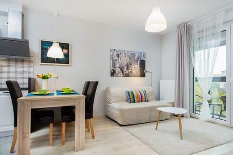 A bright and modern living room featuring a dining table with chairs, a sofa, and large windows that let in natural light. The space feels open and inviting.
