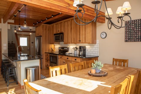 A kitchen with wooden cabinets, stainless steel appliances, a center island with bar stools, and a dining table. A chandelier hangs above the table, and a clock and word art are on the wall.