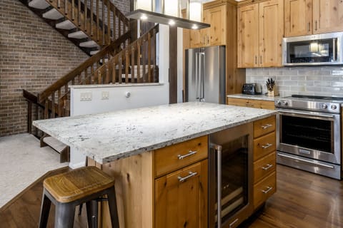Modern kitchen with wooden cabinets, a granite island countertop, stainless steel appliances, and a staircase with wooden railing in the background. A wooden stool is placed by the island.