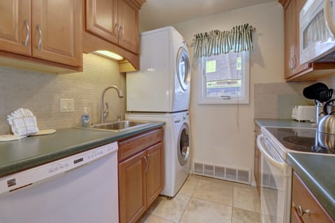 A compact kitchen with wood cabinets, a dishwasher, sink, electric stove, microwave, and stacked washer and dryer next to a small window with a green curtain.