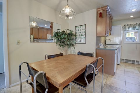 A dining area with a wooden table and four black chairs, a potted plant, wall decor, and a hanging light fixture. The space leads to a kitchen with wooden cabinets and a window with striped curtains.