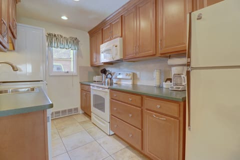 A compact kitchen with wooden cabinets, a refrigerator, stove, microwave, and coffee maker. There is a small window with a striped curtain, and green countertops next to a sink and dishwasher.