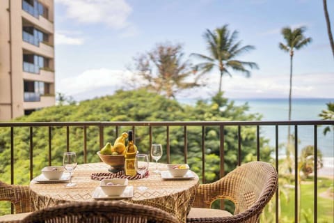 Balcony dining area with wicker table and chairs, bananas and wine glasses set against a backdrop of lush greenery, palm trees, and the ocean beyond.