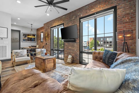 Bright and stylish living area featuring exposed brick walls, large windows with plenty of natural light, comfortable seating, a Smart TV, and a cozy modern-industrial design.