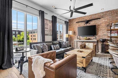 Stylish open-concept living room featuring exposed brick walls, comfortable leather seating, a large Smart TV, rustic wood accents, and a seamless flow into the fully equipped kitchen and dining area.