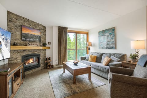 Fireplace set in a stacked stone wall with a wood mantel, flanked by a gray sofa and armchair near sliding balcony doors.