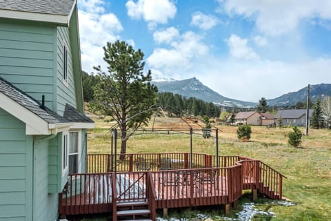 Redwood deck features metal patio furniture beneath a black-framed gazebo, overlooking open grassland and distant snow-capped peaks.