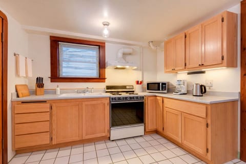 A bright, fully equipped kitchen with wood cabinetry, tiled floors, full-size stove, microwave, kettle, and ample counter space beneath a sunlit window.
