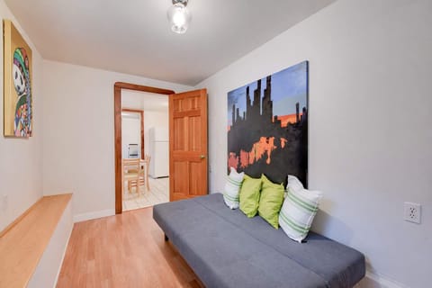 A bright sitting room with a gray sofa, colorful wall art, wood floors, built-in ledge, and an open doorway leading to the kitchen area.
