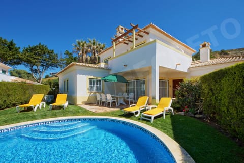 Private swimming pool at Jardim Secreto villa in Carvoeiro, with sparkling blue water, yellow sun loungers and a green parasol on the surrounding lawn, set against the backdrop of the villa and lush Algarve garden with palm trees