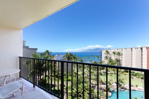 This balcony overlooks lush gardens, sparkling pool areas, swaying palms, and stunning ocean views with mountains in the distance—a tropical paradise awaits.