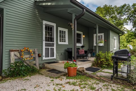 Green siding and white-trimmed windows frame the covered porch with black bench seating and a hanging flower pot.