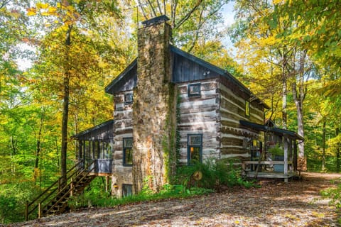Exterior view of a rustic log cabin with a tall stone chimney, surrounded by green and yellow autumn trees.